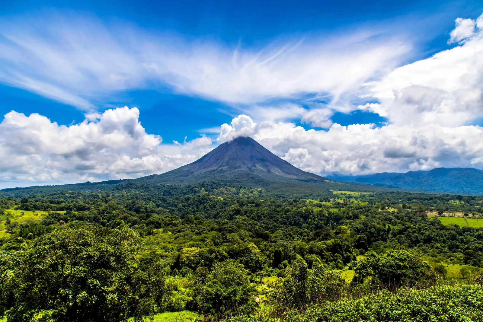 A conical volcano rising above dense green rainforest under a bright sky of towering cumulus cloud