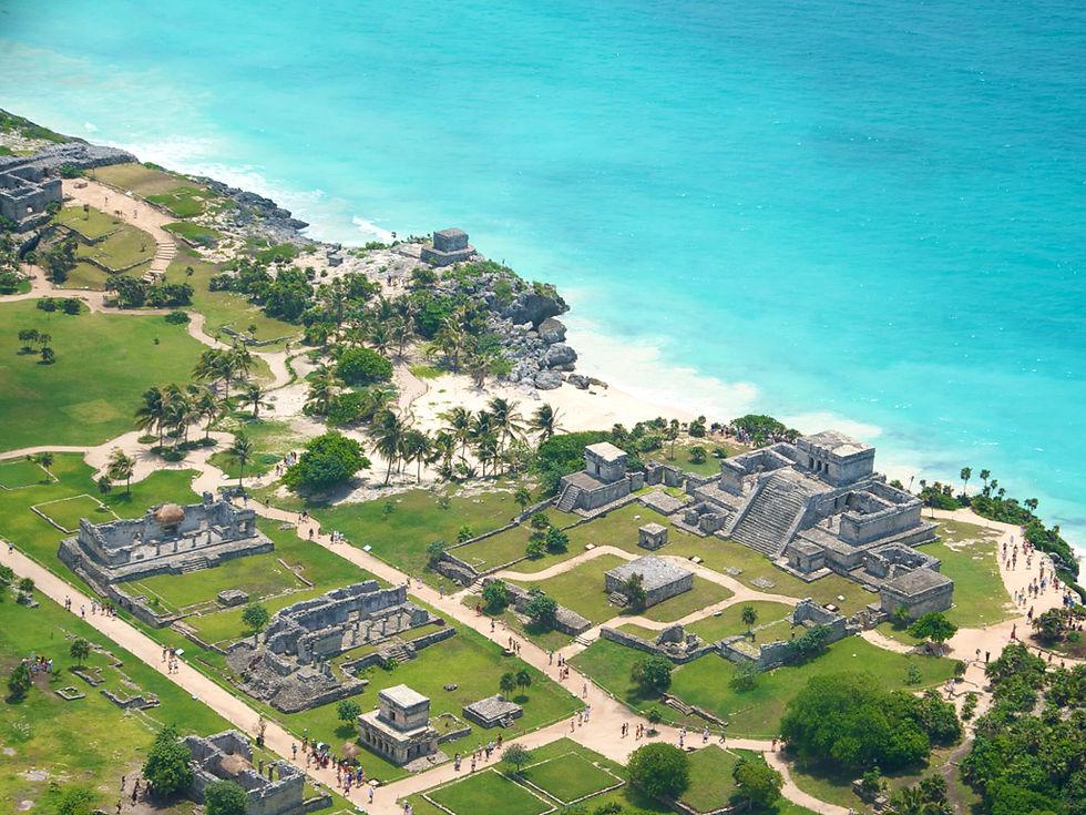 Aerial view of stone Maya ruins on a grassy clifftop above a white sand beach and turquoise Caribbean water